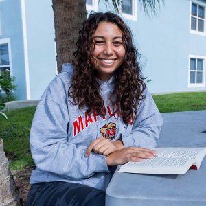 Headshot of Sofia smiling at the camera in a UMD hoodie and a book open in front of her.
